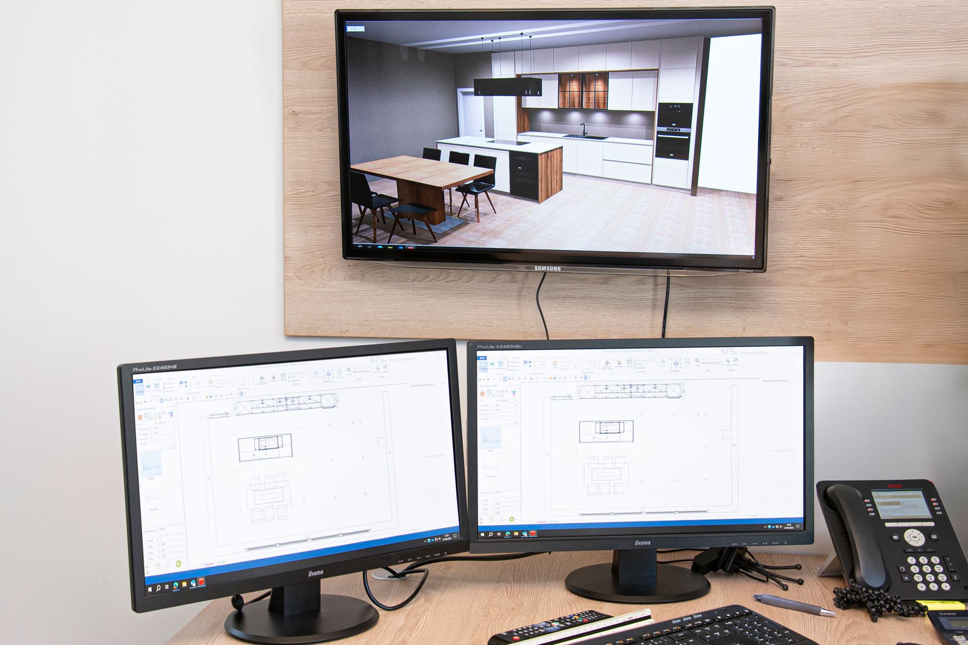 Auf drei Monitoren werden eine Küchenplanungssoftware und ein 3D-Küchenrender angezeigt; auf dem Schreibtisch daneben liegt ein Telefon.