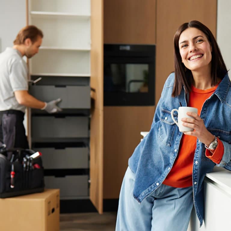Eine lächelnde Frau mit einer Tasse lehnt sich an einen Tresen, während ein Arbeiter in einer modernen Küche Schränke repariert.
