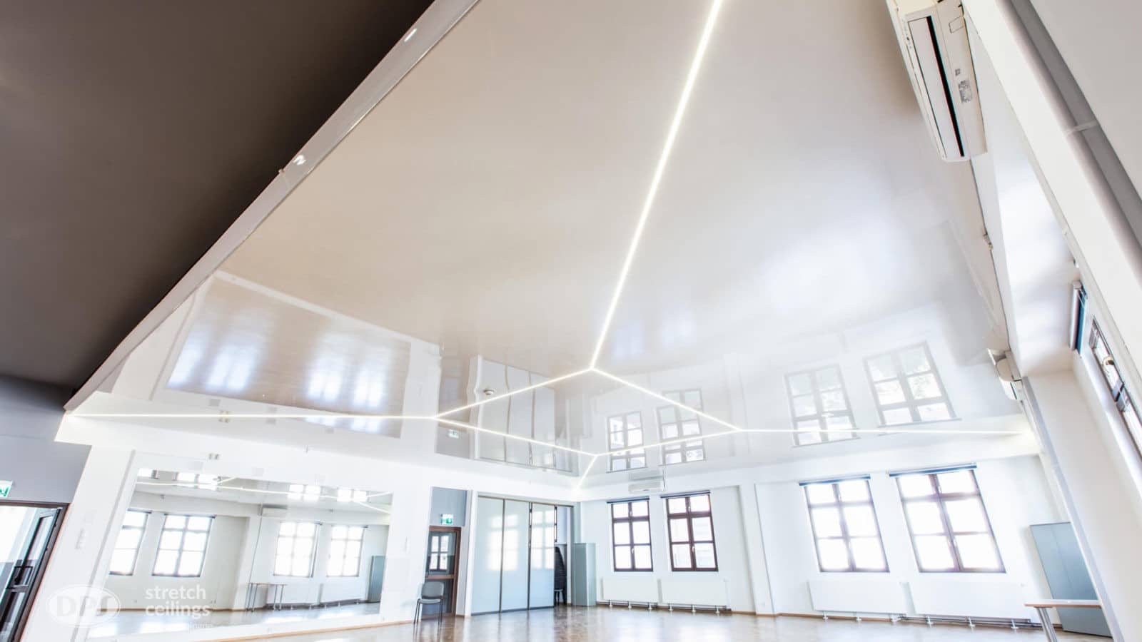 Ein moderner, geräumiger Raum mit hoher, glänzender Decke und eckigen LED-Leuchten, mit großen Fenstern und weißen Wänden.