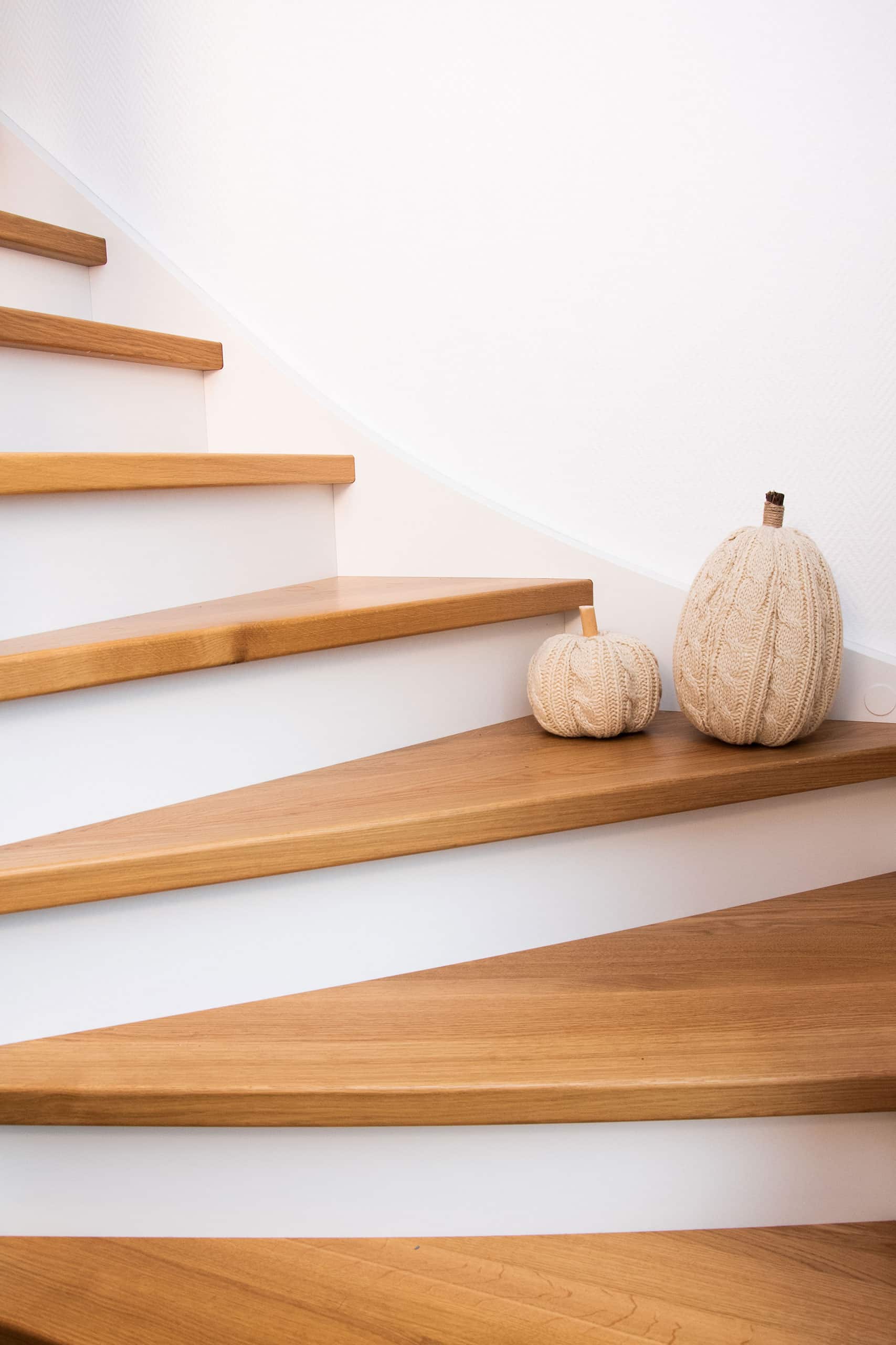 Zwei mit Strick überzogene Zierkürbisse stehen auf einer modernen Holztreppe vor einer weißen Wand.