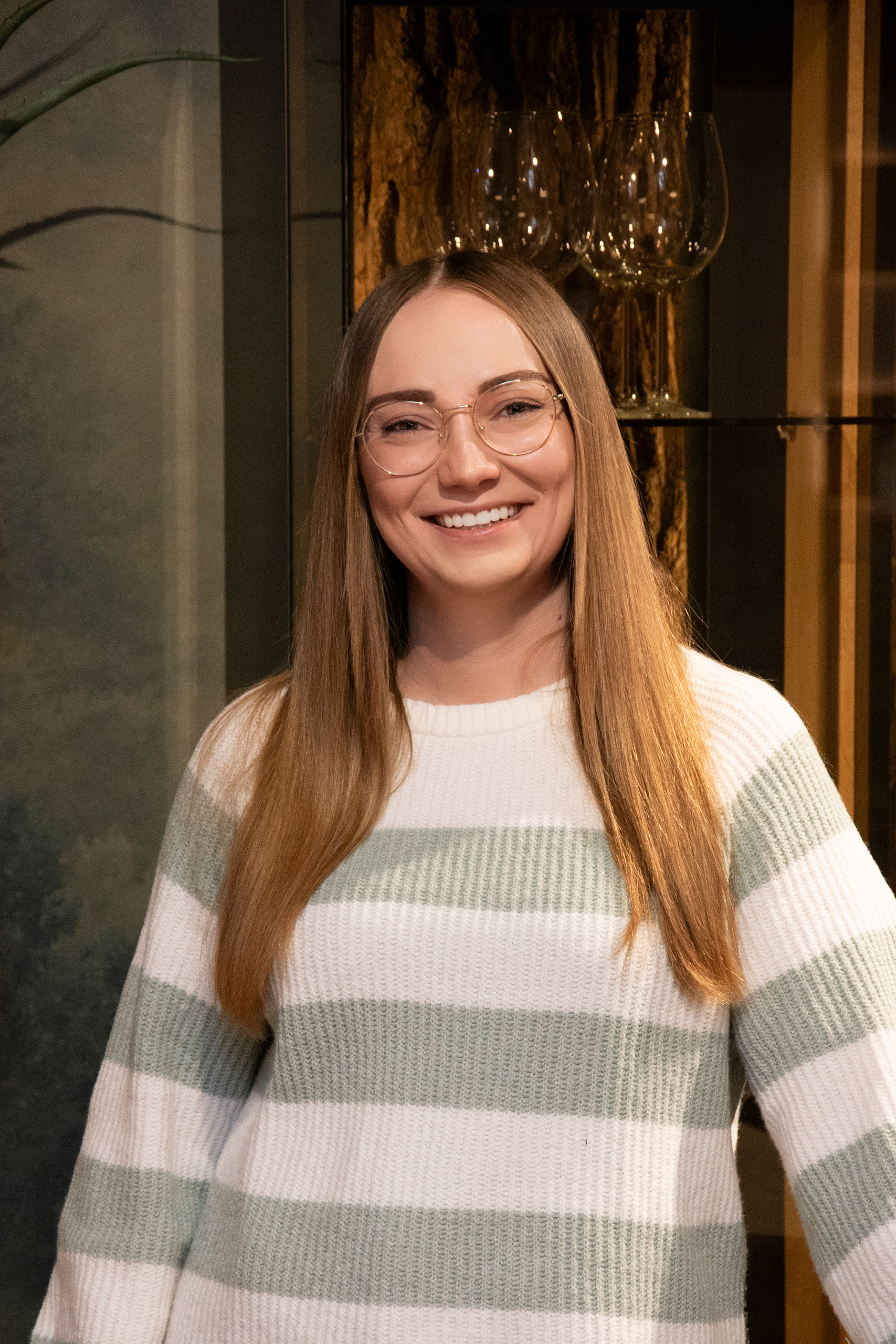 Eine Frau mit langen glatten Haaren und Brille lächelt, sie trägt einen weiß-hellgrün gestreiften Pullover.