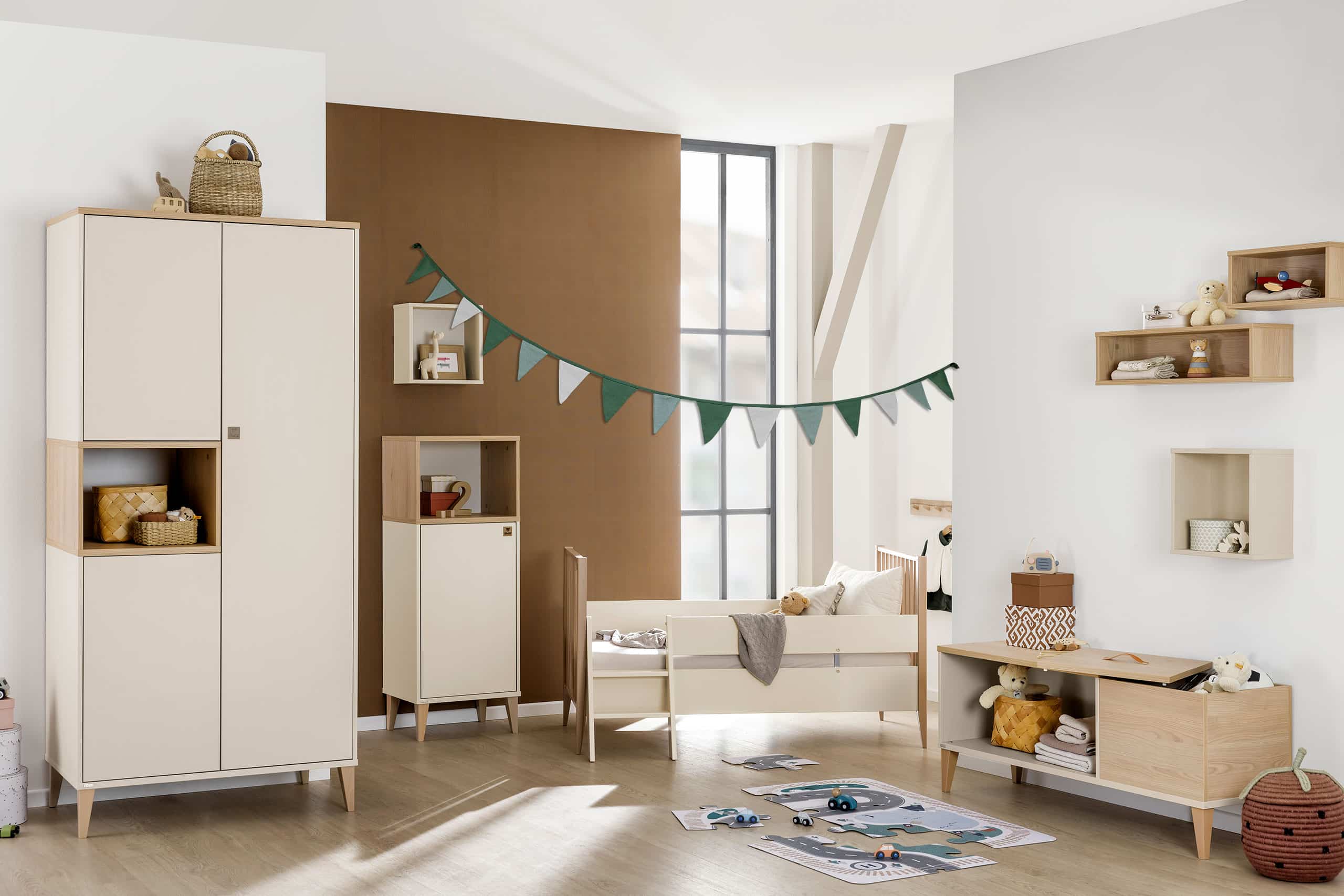 Modernes, gemütliches Kinderzimmer mit beigen Möbeln, einem kleinen Bett, Wandregalen, Spielzeug und einem grünen, dreieckigen Banner, das quer durch den Raum hängt.