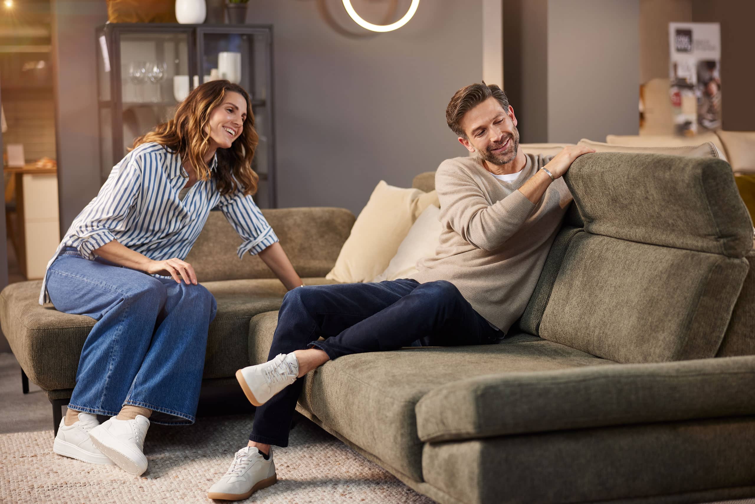 Ein lächelnder Mann und eine lächelnde Frau entspannen sich und unterhalten sich, während sie auf einem modernen olivgrünen Sektionssofa in einem Wohnzimmer sitzen.