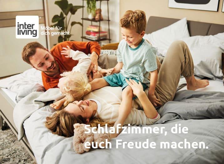 Eine glückliche vierköpfige Familie lacht und spielt zusammen auf einem Bett in einem hellen, gemütlichen Schlafzimmer.