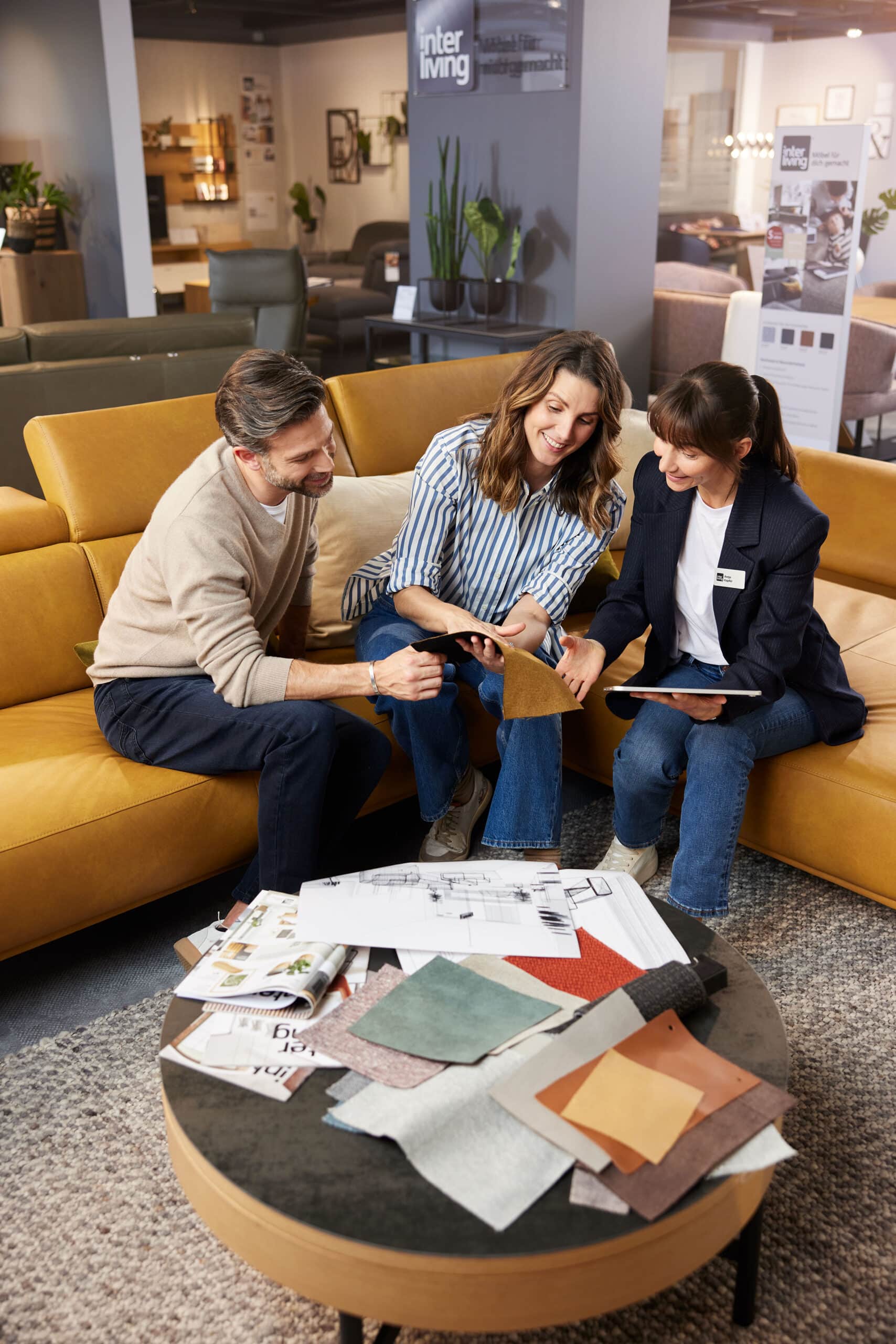 Ein Ehepaar wählt mit einem Verkaufsberater in einem Möbelhaus auf einem gelben Sofa sitzend Stoffmuster aus.