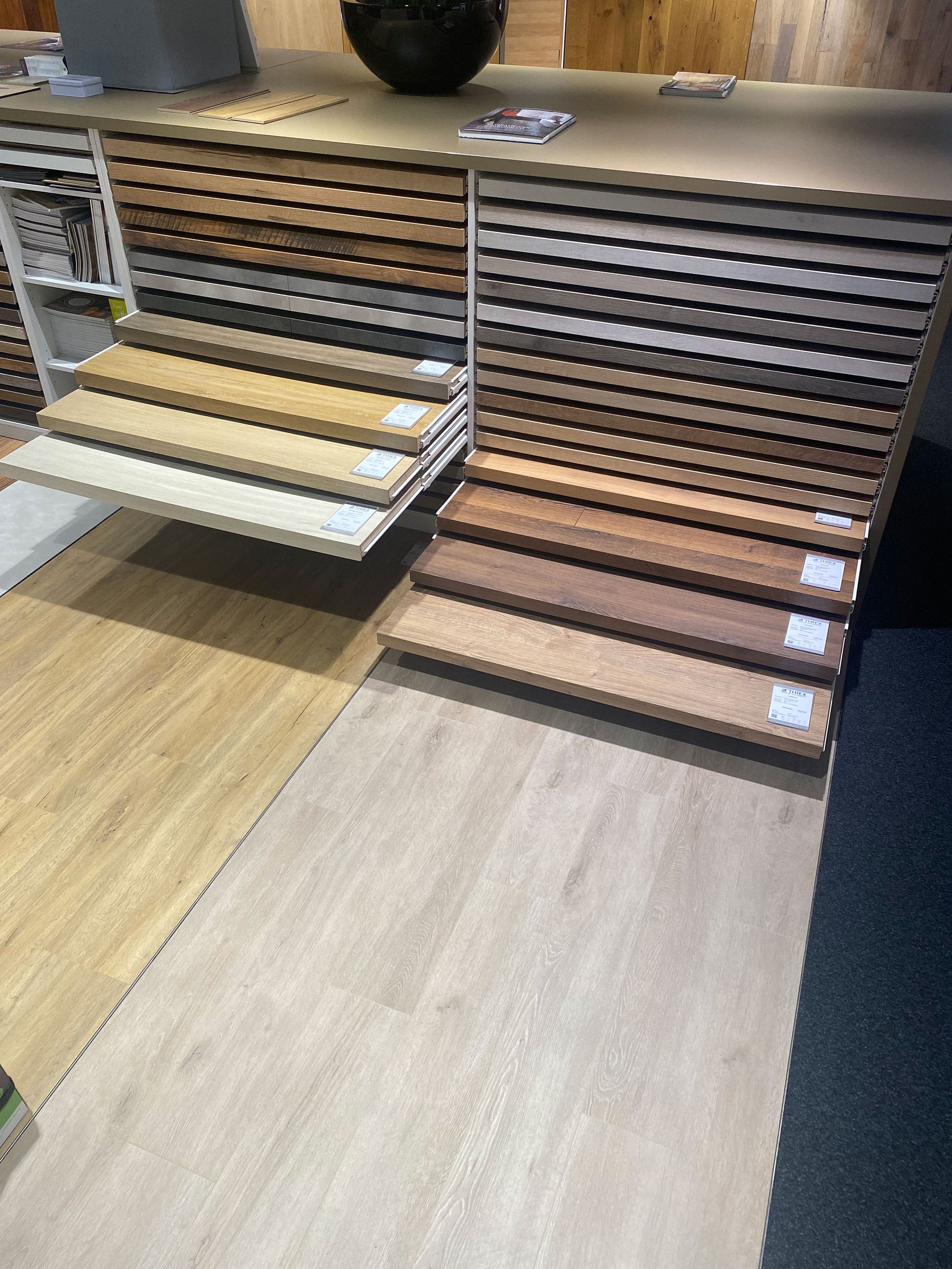 Auslage von Holzbodenmustern in verschiedenen Farbtönen, angeordnet auf ausziehbaren Regalen in einem Ausstellungsraum.