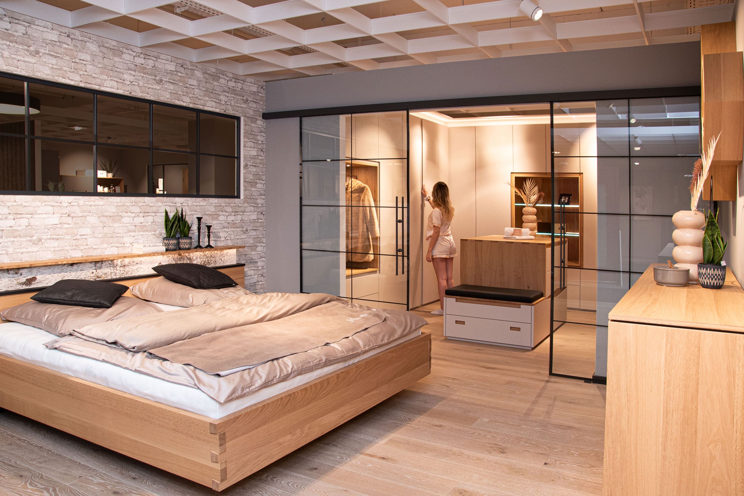 Modernes Schlafzimmer mit Holzmöbeln, großem Bett und einem begehbaren Kleiderschrank aus Glas, in dem eine Frau steht.