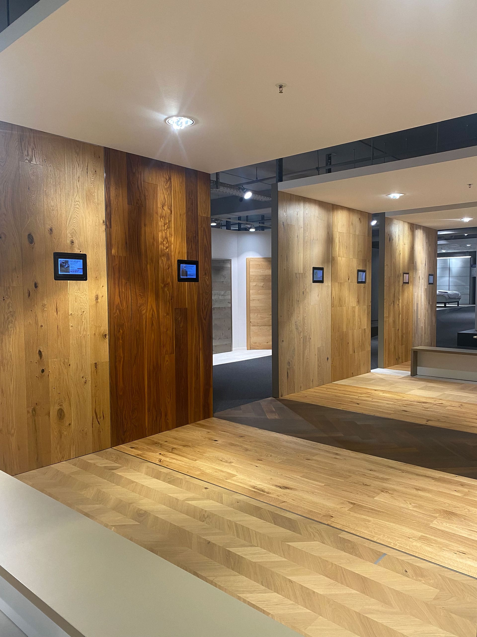 Drei hölzerne Wandpaneele mit kleinen Bildschirmen in einem modernen, gut beleuchteten Ausstellungsraum mit Holzfußboden.