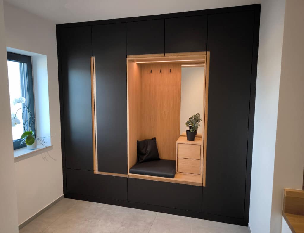 Moderner Einbauschrank in Schwarz und Holz mit einer gemütlichen Bank, Kissen, Schubladen, Haken und einer Pflanze in der Nähe eines Fensters.