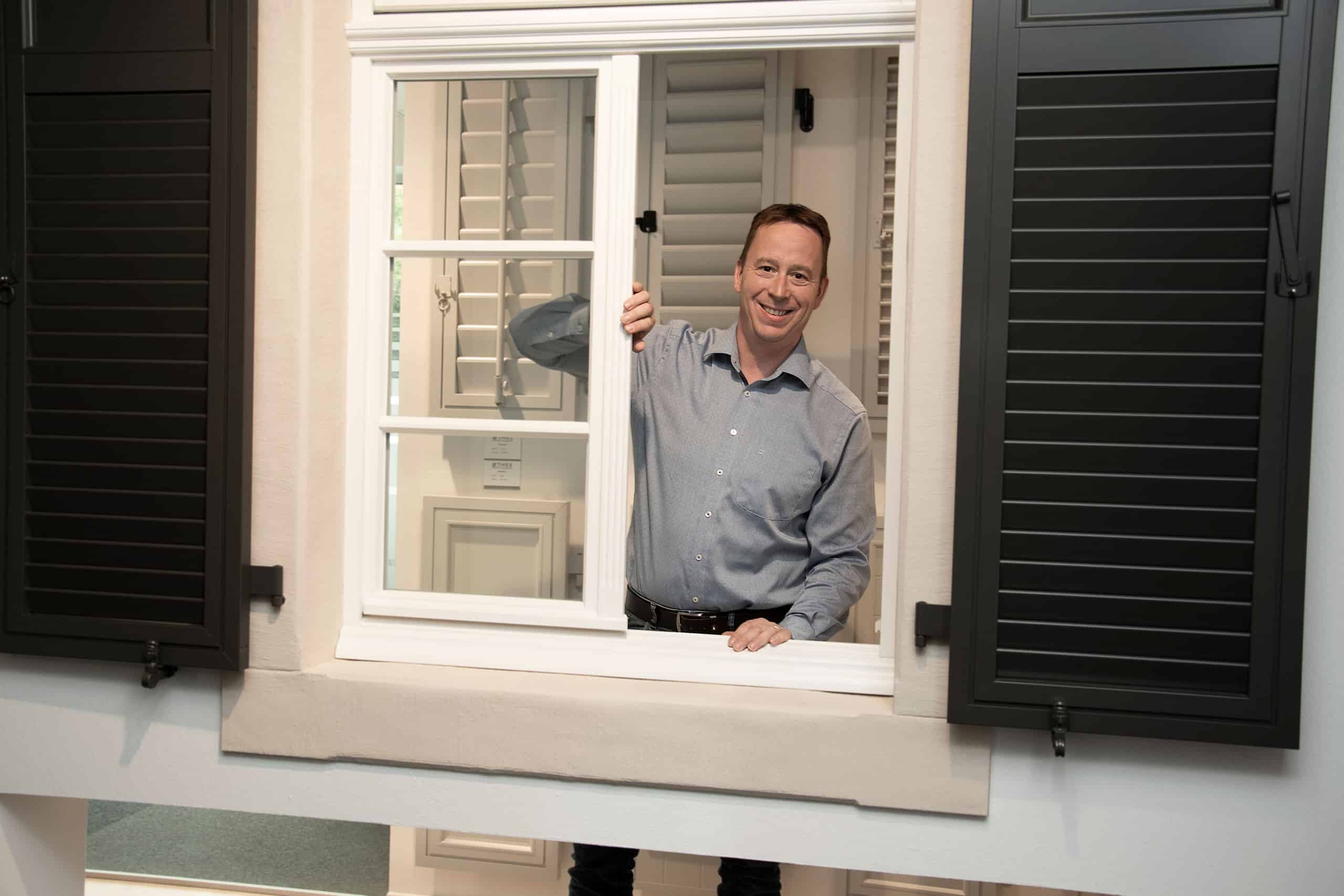 Ein Mann in einem hellblauen Hemd lächelt, während er ein weiß gerahmtes Fenster mit schwarzen Fensterläden öffnet.