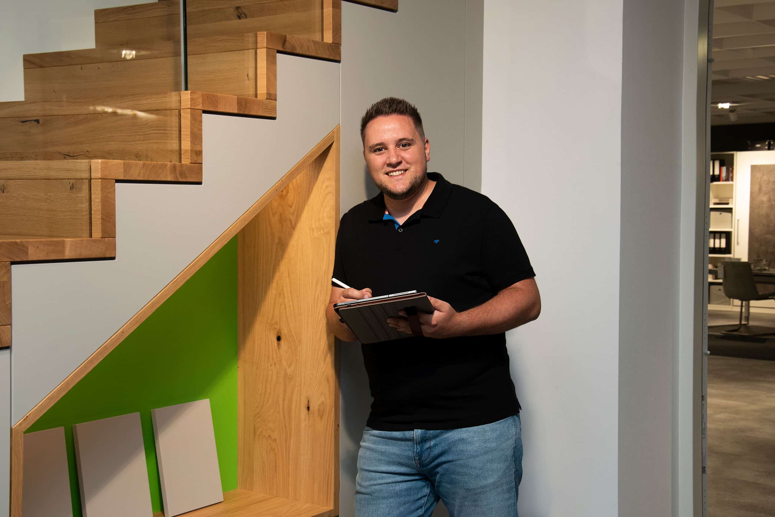 Ein Mann in schwarzem Hemd und Jeans lächelt, während er unter einer modernen Holztreppe in einem Innenraum auf ein Tablet schreibt.
