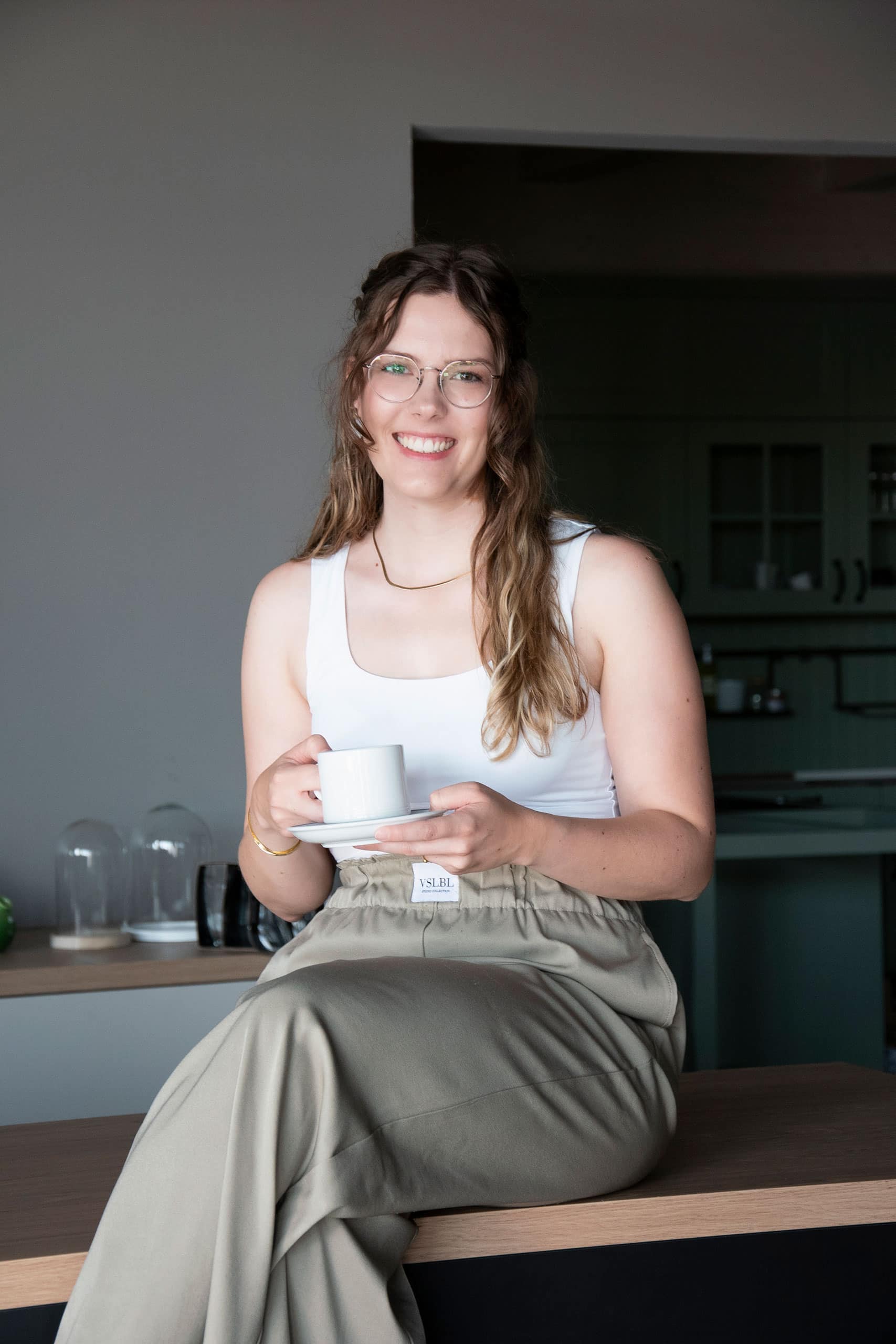 Frau mit langen Haaren und Brille sitzt mit einer weißen Tasse in der Hand lächelnd in einer modernen Küche.