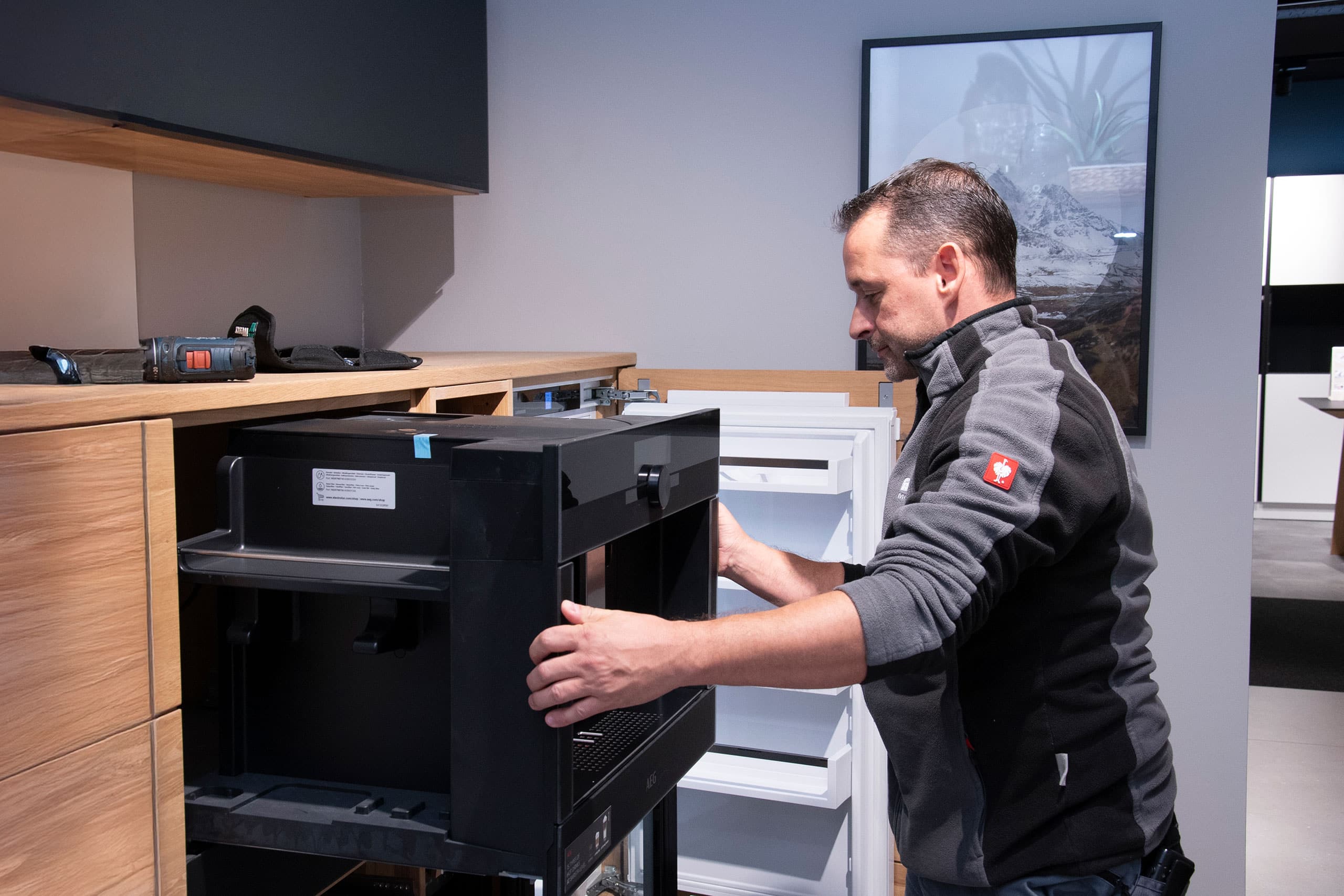Ein Mann baut ein schwarzes Küchengerät in einen Holzschrank neben einem weißen Kühlschrank ein.