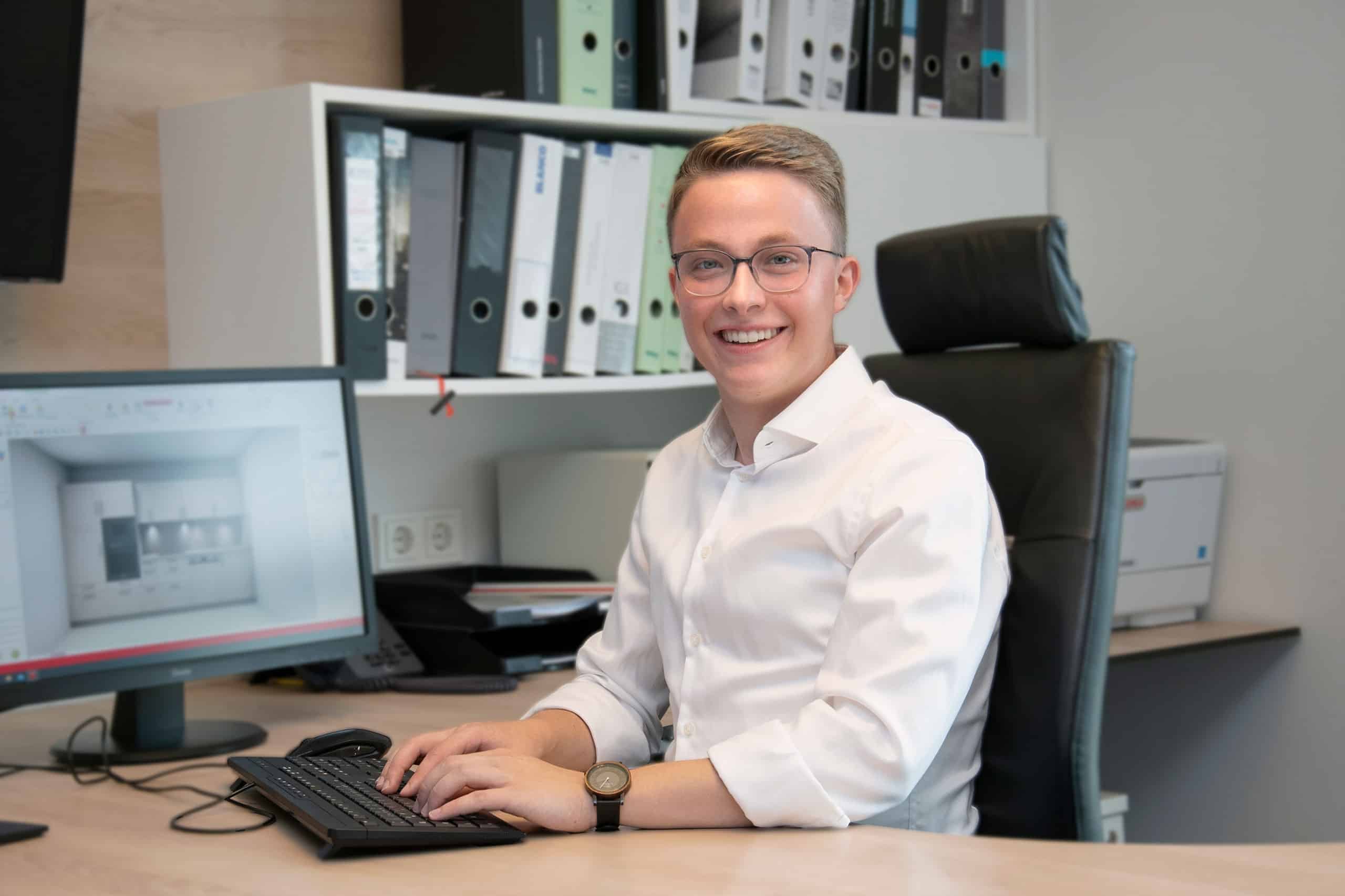Ein lächelnder junger Mann mit Brille arbeitet an einem Schreibtisch mit einem Computer und Büroakten im Hintergrund.