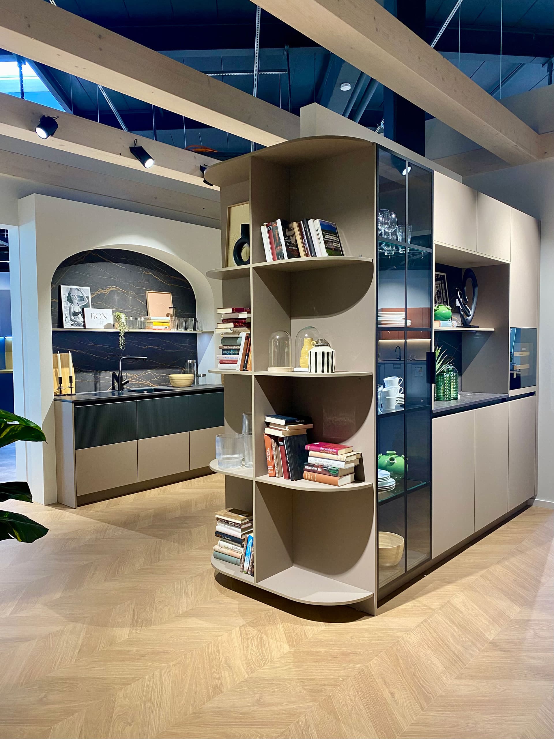 Moderne Küche mit offenen Regalen für Bücher und Dekoration, hellem Holzboden und dunklen Arbeitsplatten im Hintergrund.