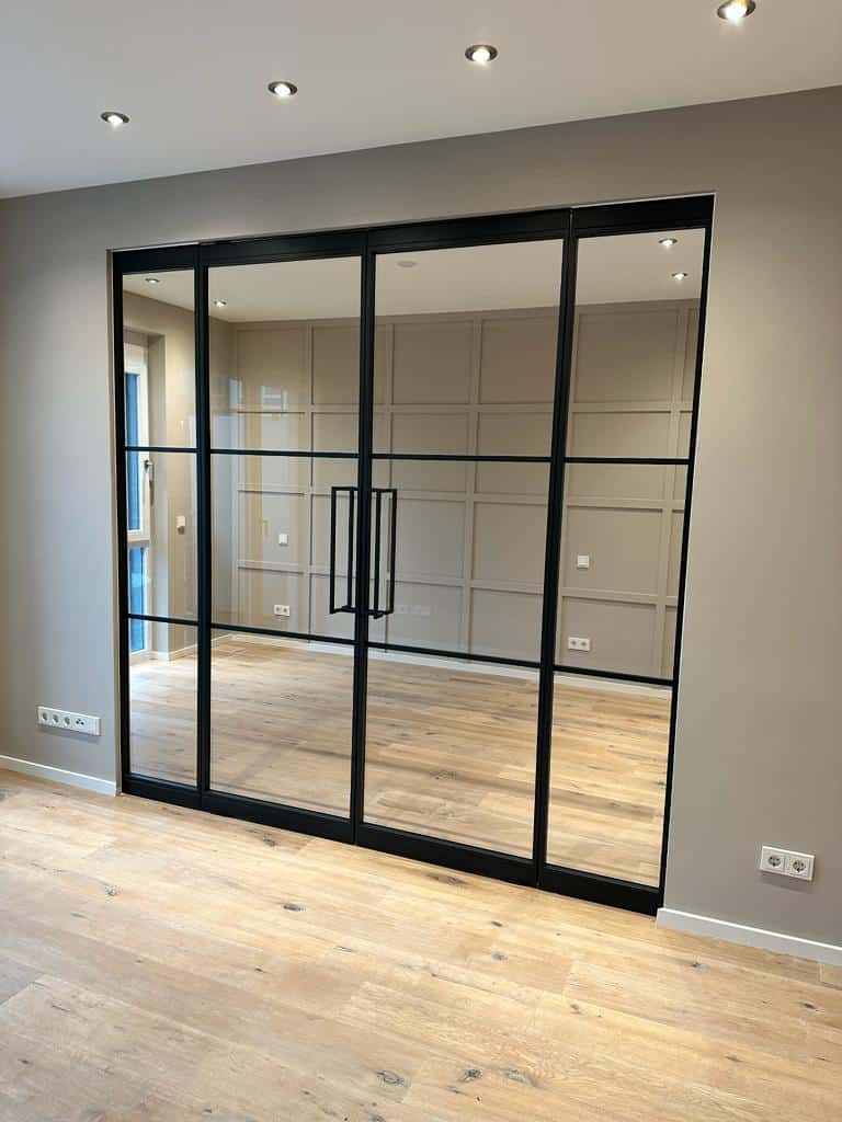 Große Glasschiebetüren mit schwarzen Rahmen trennen zwei moderne Räume mit Holzfußboden und beige getäfelten Wänden.