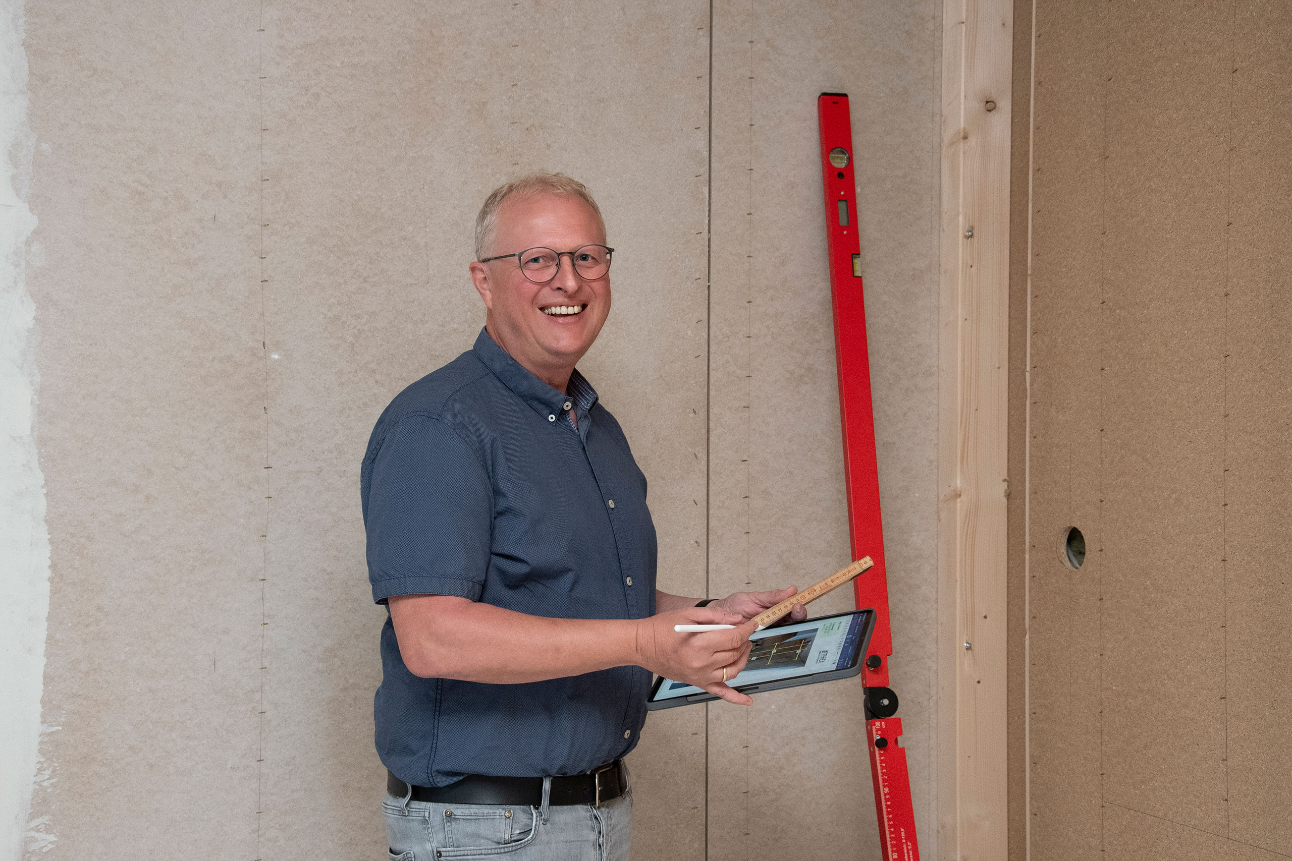 Lächelnder Mann mit Klemmbrett und Bleistift, der an einer Wand mit einem roten Nivelliergerät steht.