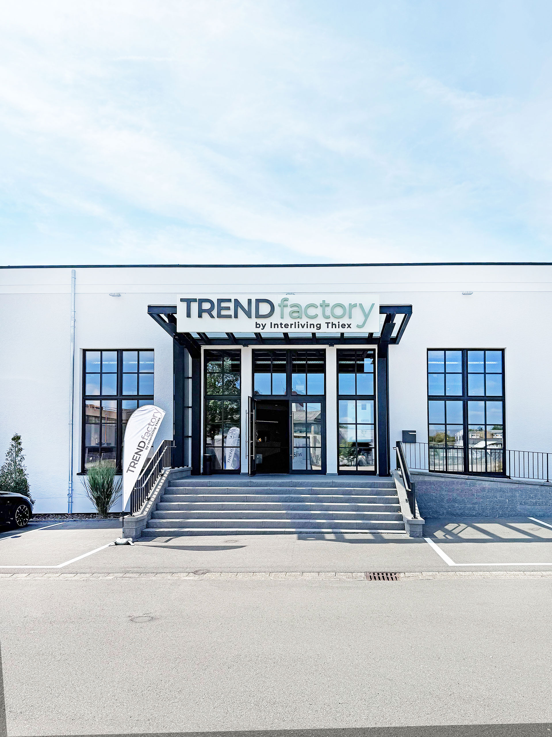 Modernes weißes Gebäude mit großen Fenstern und einem Schild mit der Aufschrift TRENDfactory by Interliving Thiex über dem Eingang.