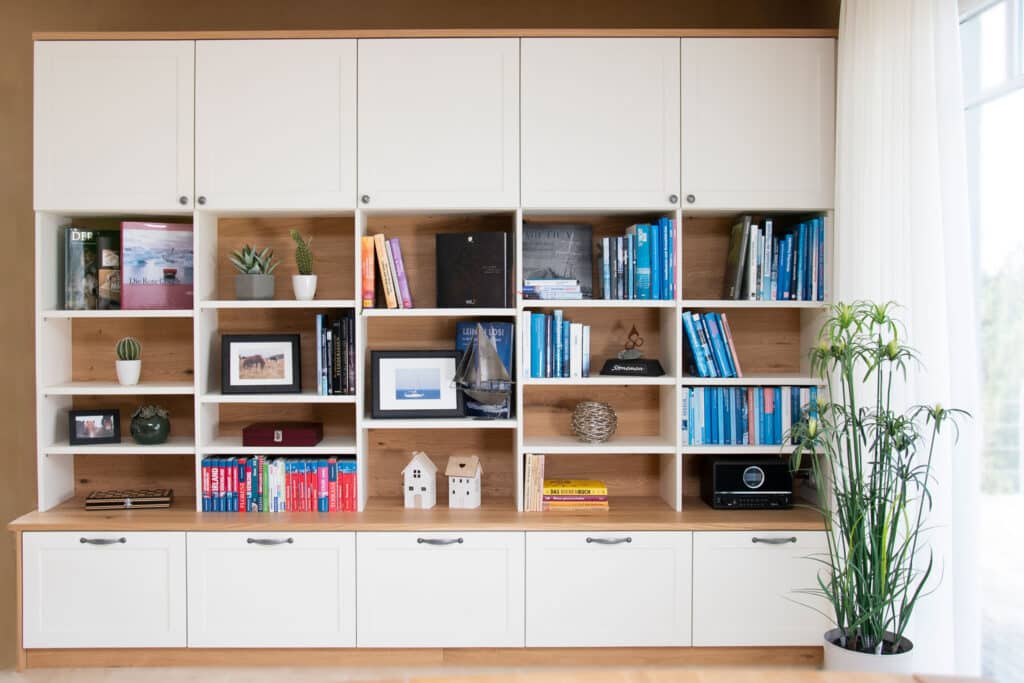 Ein weißes Bücherregal mit Büchern, gerahmten Fotos, Pflanzen, Dekoartikeln und Schränken, daneben eine hohe Topfpflanze.