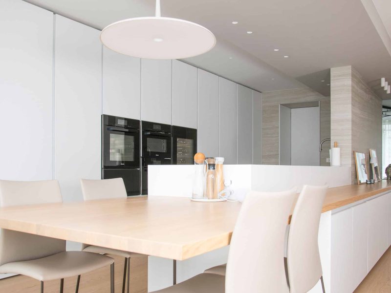 Minimalistische moderne Küche mit hellem Holztisch, weißen Stühlen und schwarzen Einbaugeräten an einer weißen Wand.