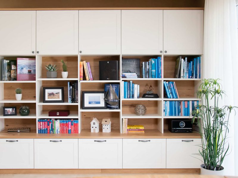 Ein weißes Bücherregal mit Büchern, gerahmten Fotos, Pflanzen, Dekoartikeln und Schränken, daneben eine hohe Topfpflanze.